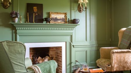 Living room with fireplace and walls painted in Calke Green by Farrow & Ball, two armchairs, and a mantelpiece with antique paintings, vases and candlesticks