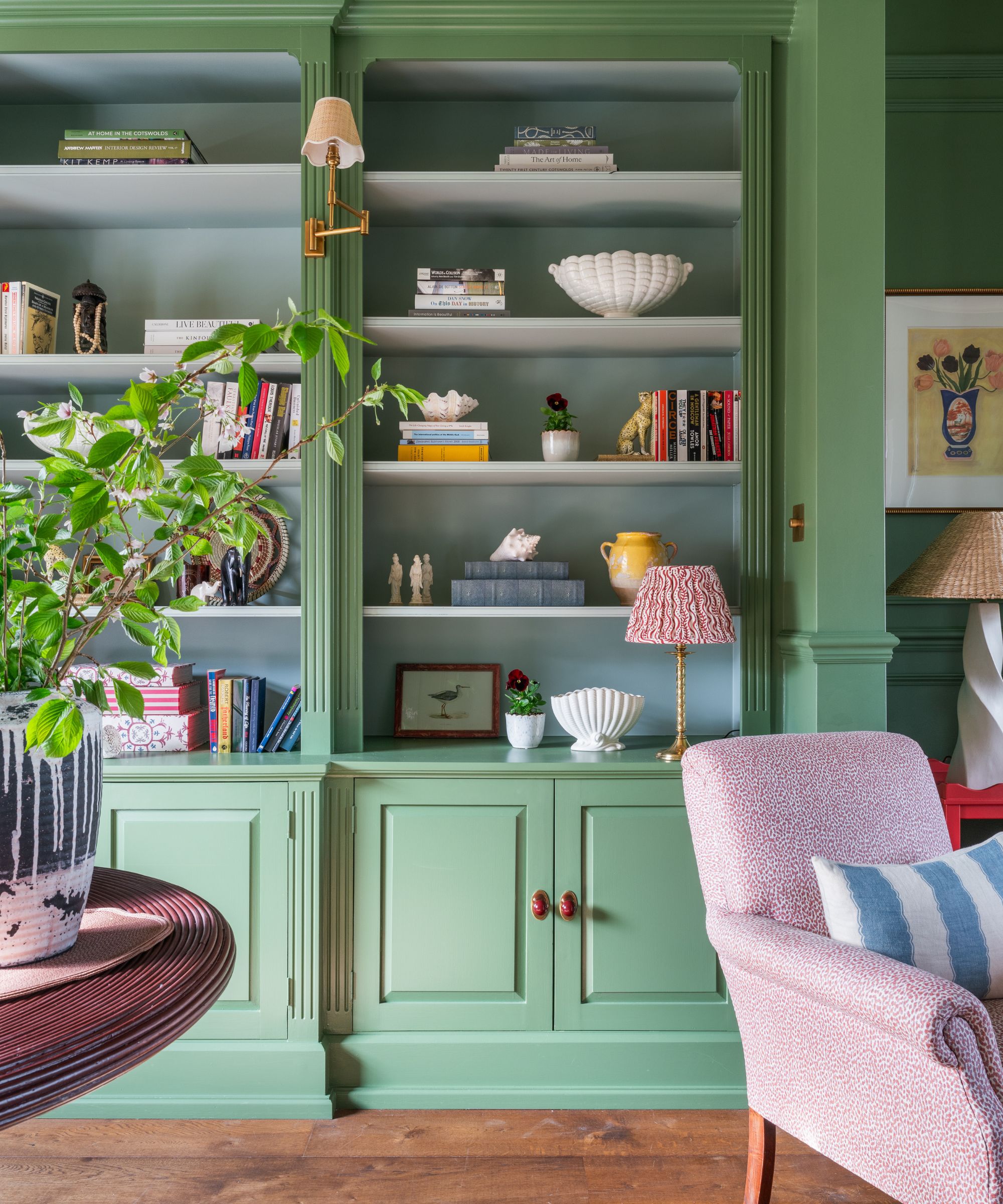 Living room with a built-in bookcase painted in Farrow &amp;amp; Ball's Calke green, decorated with books, ceramics, and trinkets, a pink armchair with a striped blue cushion, and a dark wood round table
