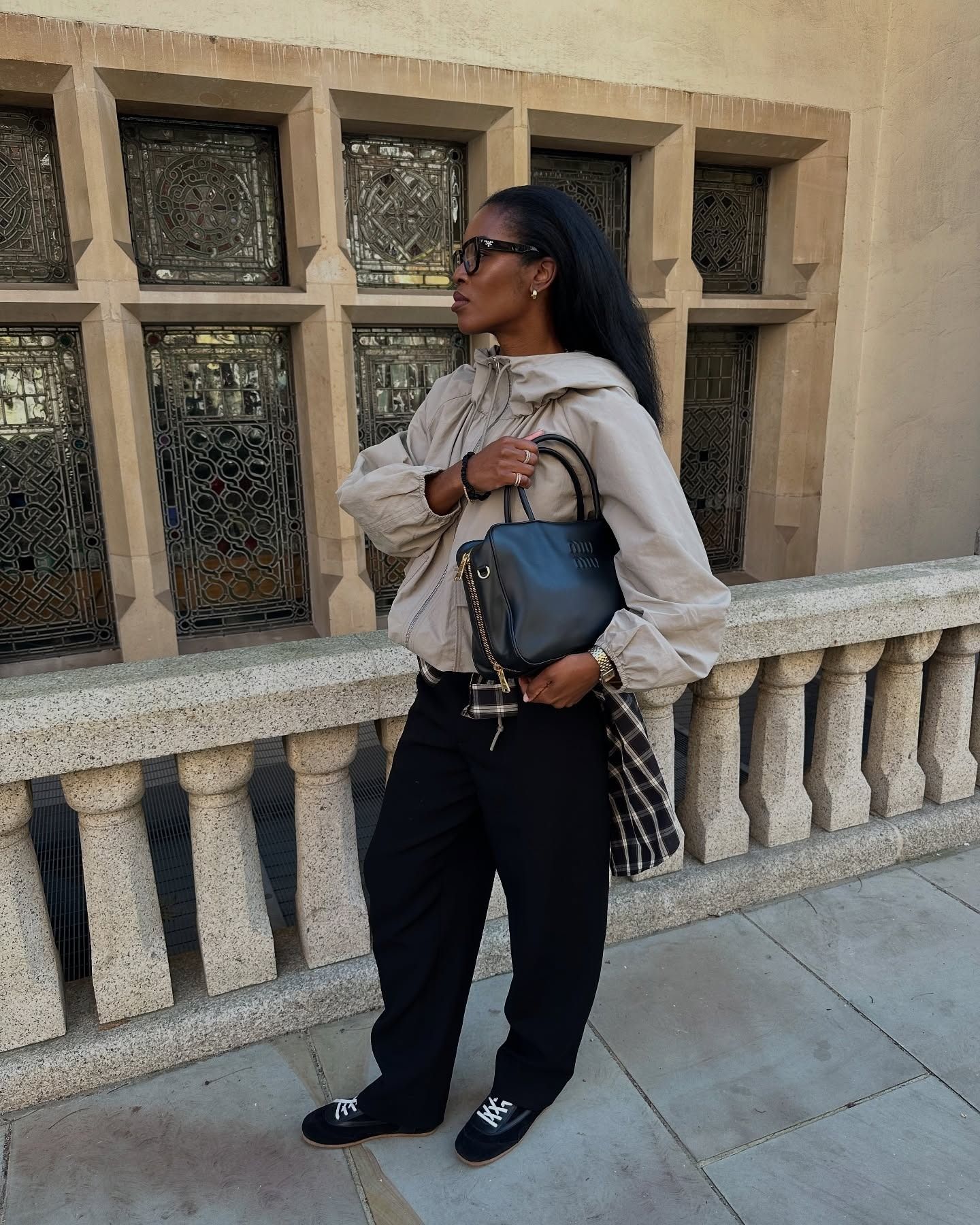 Woman wearing stone windbreaker, with black pants and black sneakers, carrying black handbag.