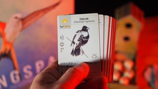 A hand holding a selection of Wingspan cards, with the box and dicetower in the background