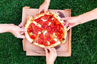 A pizza in it's box against a green grass background. Several hands are reaching for the pizza