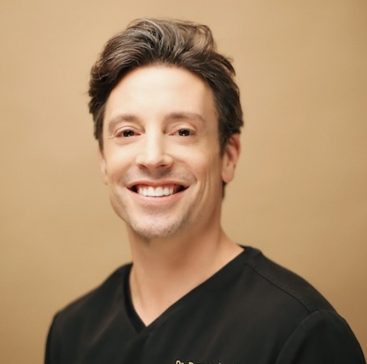 a headshot of a white man in black scrubs smiling at the camera