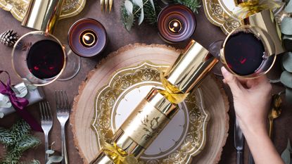 A gold Christmas cracker on a plate, surrounded by drink glasses and Chirstmas table decorations
