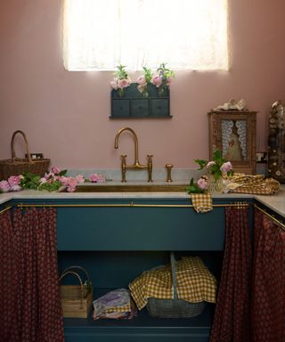 pink laundry room with blue cabinets with cafe curtain, white countertops and brass taps