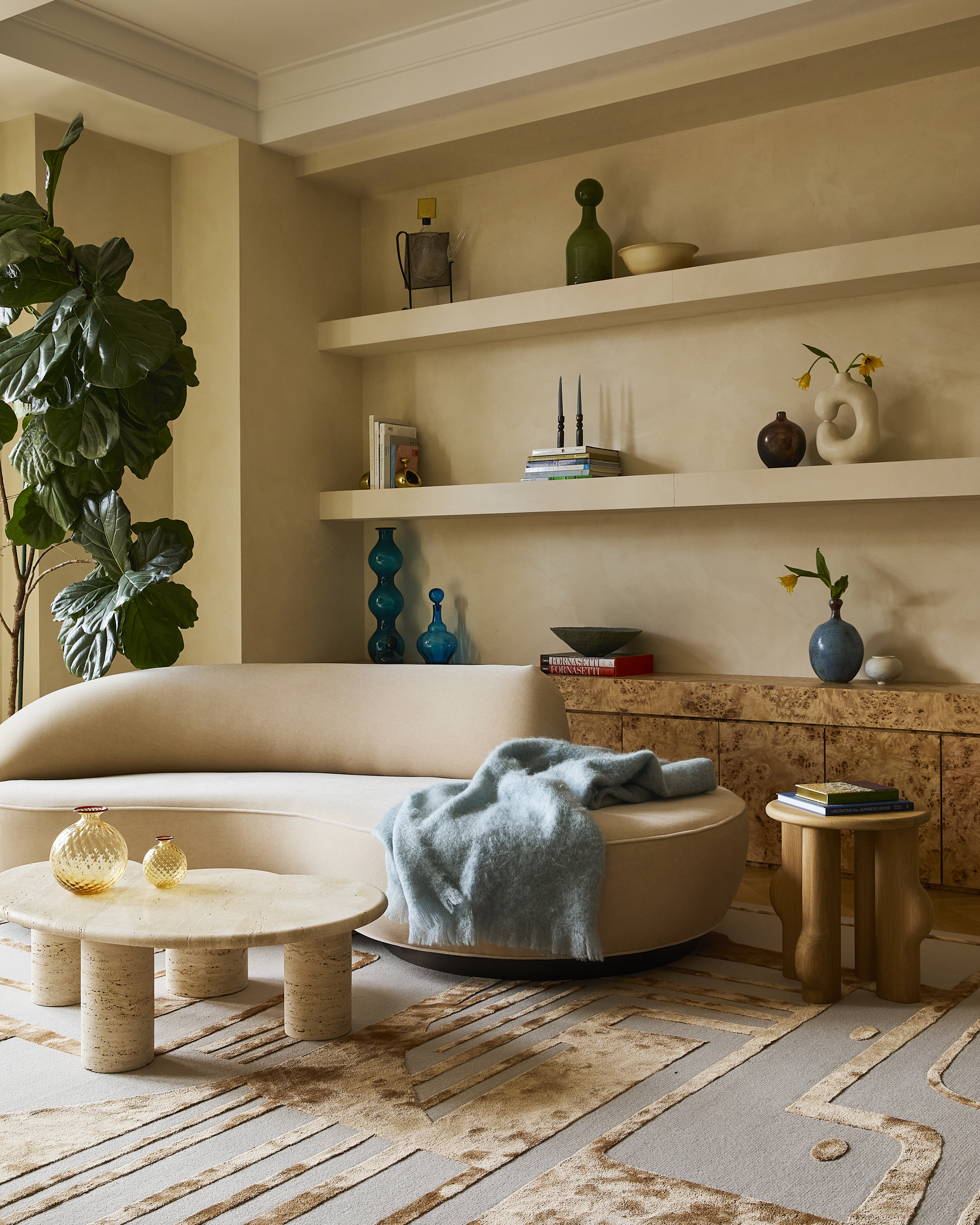neutral living room with floating shelves, limewash walls, curved beige sofa with blue throw draped over, travertine round coffee table, blue and gold tufted rug, burl console, fiddle leaf potted tree, and side table styled with books