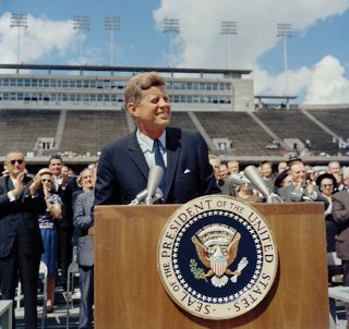 a smiling man in a suit stands at a wooden lectern in a mostly empty football stadium.