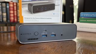 A gray HyperDrive Next docking station with multiple ports displayed on a wooden desk, surrounded by colorful books and a framed image.