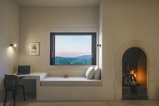 A room featuring a bed made of concrete base with an extension to a work table with a laptop placed on it. A white frame on the wall next to the wide window with views of the mountain
