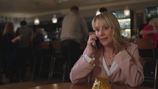 Kellie Shirley as nurse Sophia Peters sitting in a pub wearing a white blouse and pink coat, on the phone and looking upset.