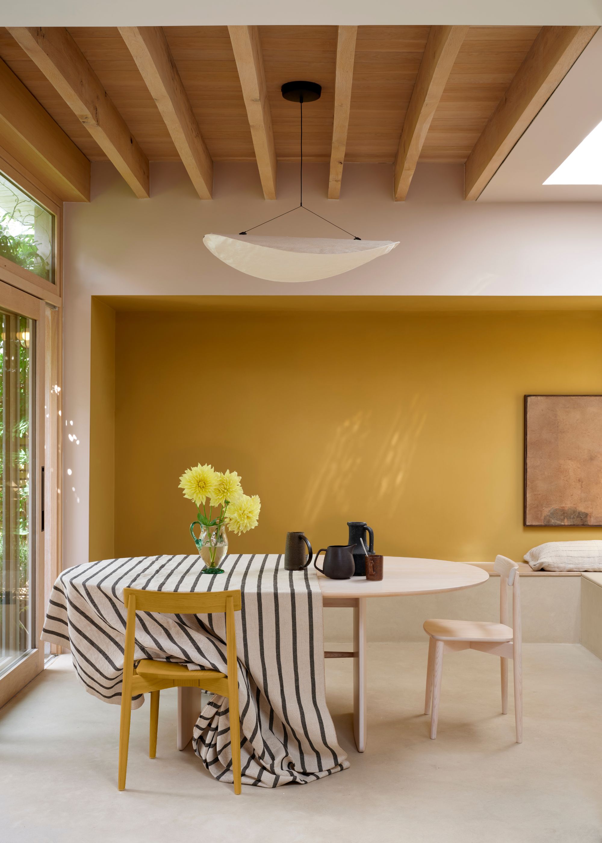 A light and airy dining room with a bold yellow accent wall, a wooden dining table and chairs, with a striped tablecloth.