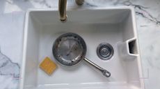 Ceramic sink with marble surround and brass tap with burnt stainless steel in sink next to a sponge to question the methods for how to clean a burnt pan