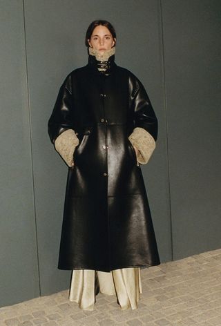 one of the best jacket coat brands, Nanushka is shown in an image of a model wearing a long black leather funnel neck jacket over a satin dress