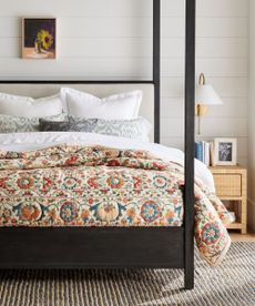 Front view of four-poster black wooden bed, white bedding with patterned throw and pillow, gray stroped rug and wicker nightstand