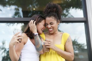 Two millennial women, one Caucasian with shoulder length hair and the other mixed race with curly hair, laugh while they hug each other outdoor near a building, wearing casual summer clothing.