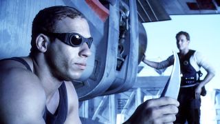 Still from the movie series The Chronicles of Riddick. Here we see Riddick (bald man with dark goggles) sitting staring intently at the sharp blade in his hands. In the background is a man, wearing dark armor, watching Riddick.