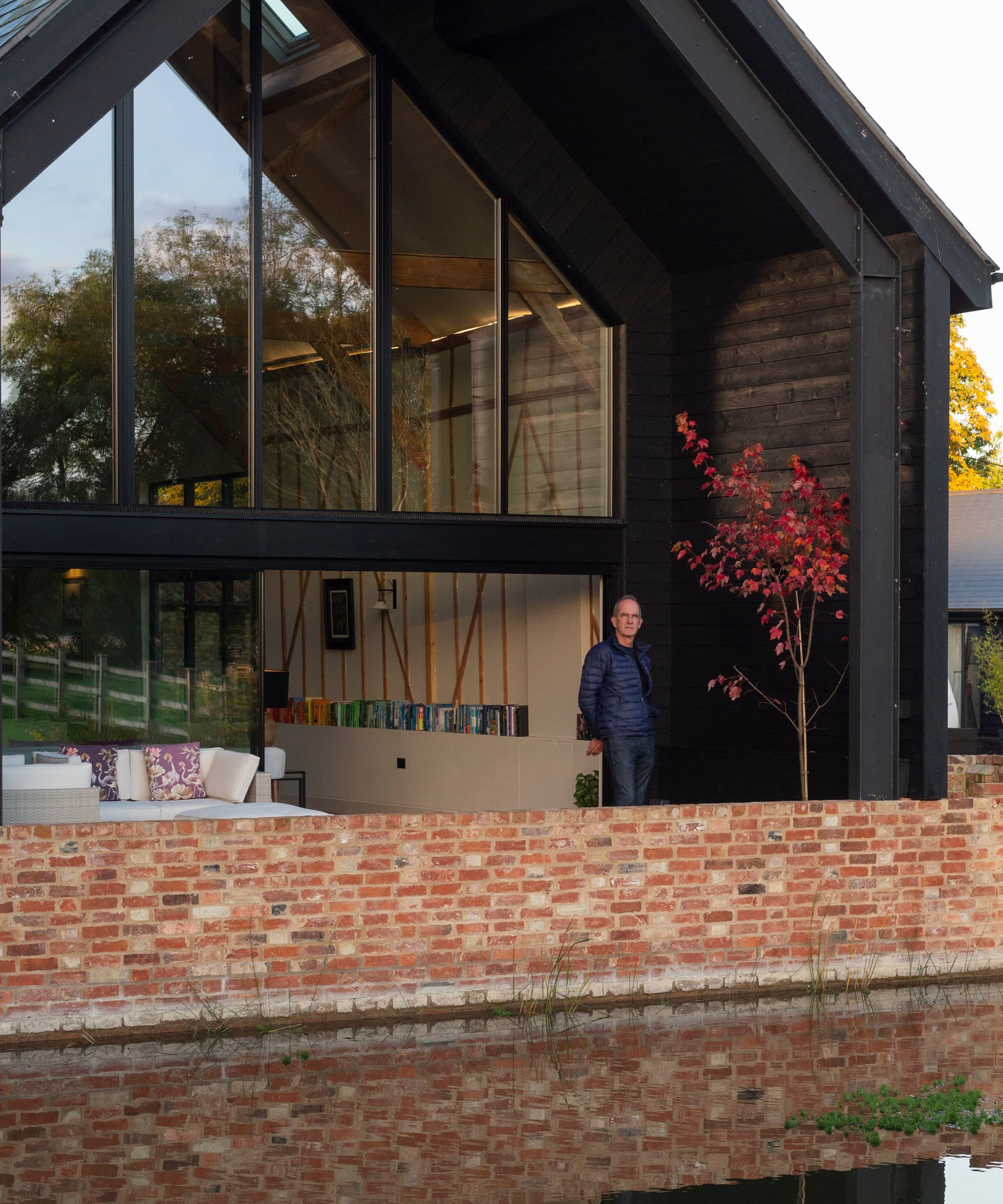 Kevin McCloud standing in front of a barn conversion with black cladding and next to a brick wall next to a river