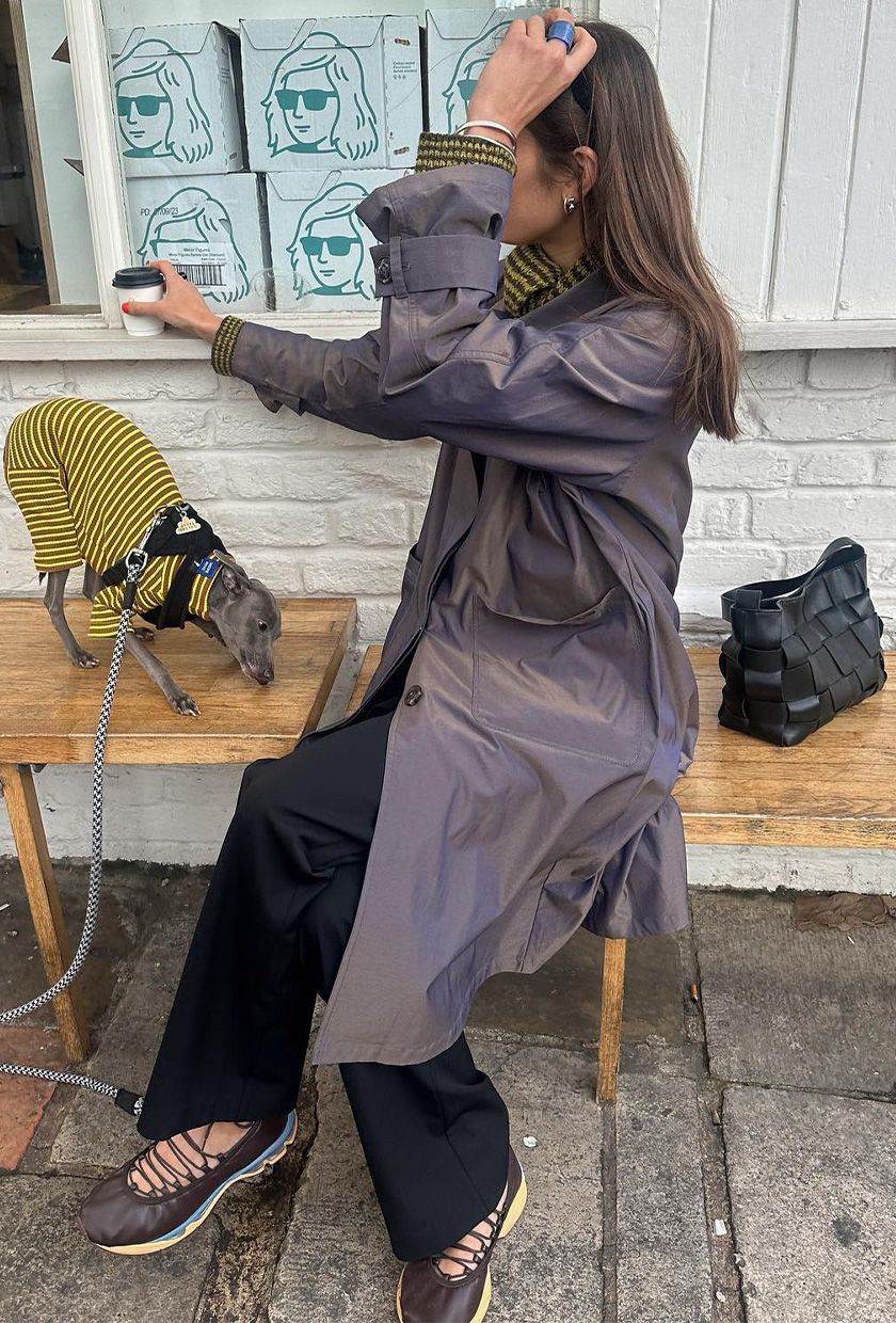 The best ballet sneakers are shown in a photo of a woman sitting on a bench outside a coffee shop with her dog wearing a gray trench coat with black trousers, a stripped yellow and black sweater, and brown multi-color lace-up ballerina sneakers