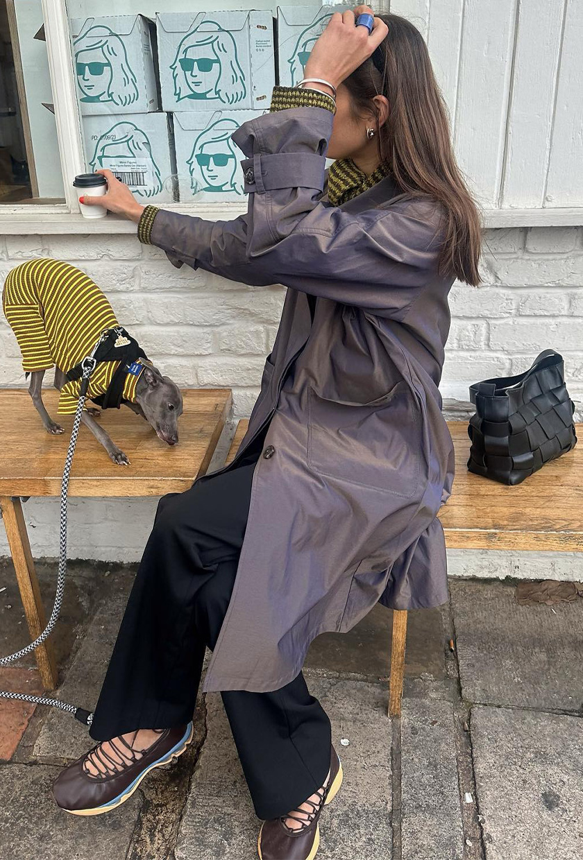 The best ballet sneakers are shown in a photo of a woman sitting on a bench outside a coffee shop with her dog wearing a gray trench coat with black trousers, a stripped yellow and black sweater, and brown multi-color lace-up ballerina sneakers