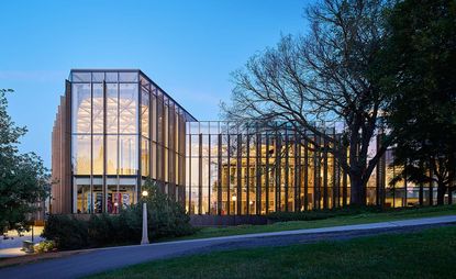 A street level view of the front of the National Arts Centre which is predominantly made of glass panel and has an illuminated interior. 