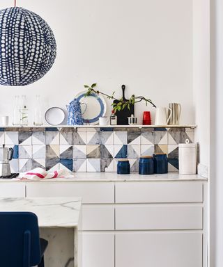 A blue and white kitchen color scheme with colorful tiles and shelf with accessories above white handless cabinets.