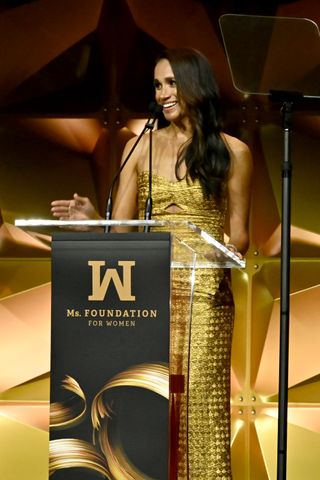 Meghan Markle speaking onstage in a gold dress