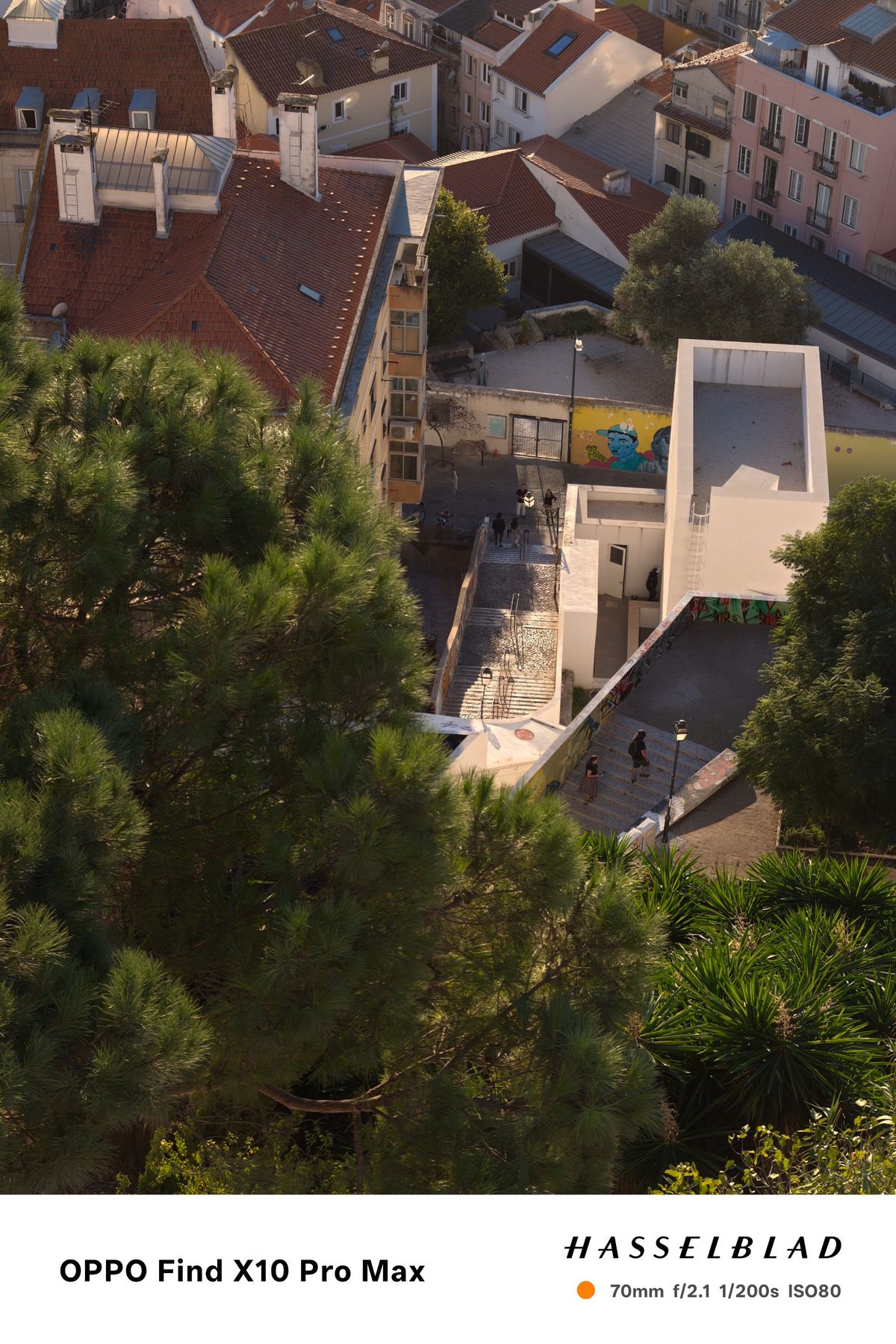 Elevated view over Lisbon rooftops, trees and a modern white building in warm evening light.