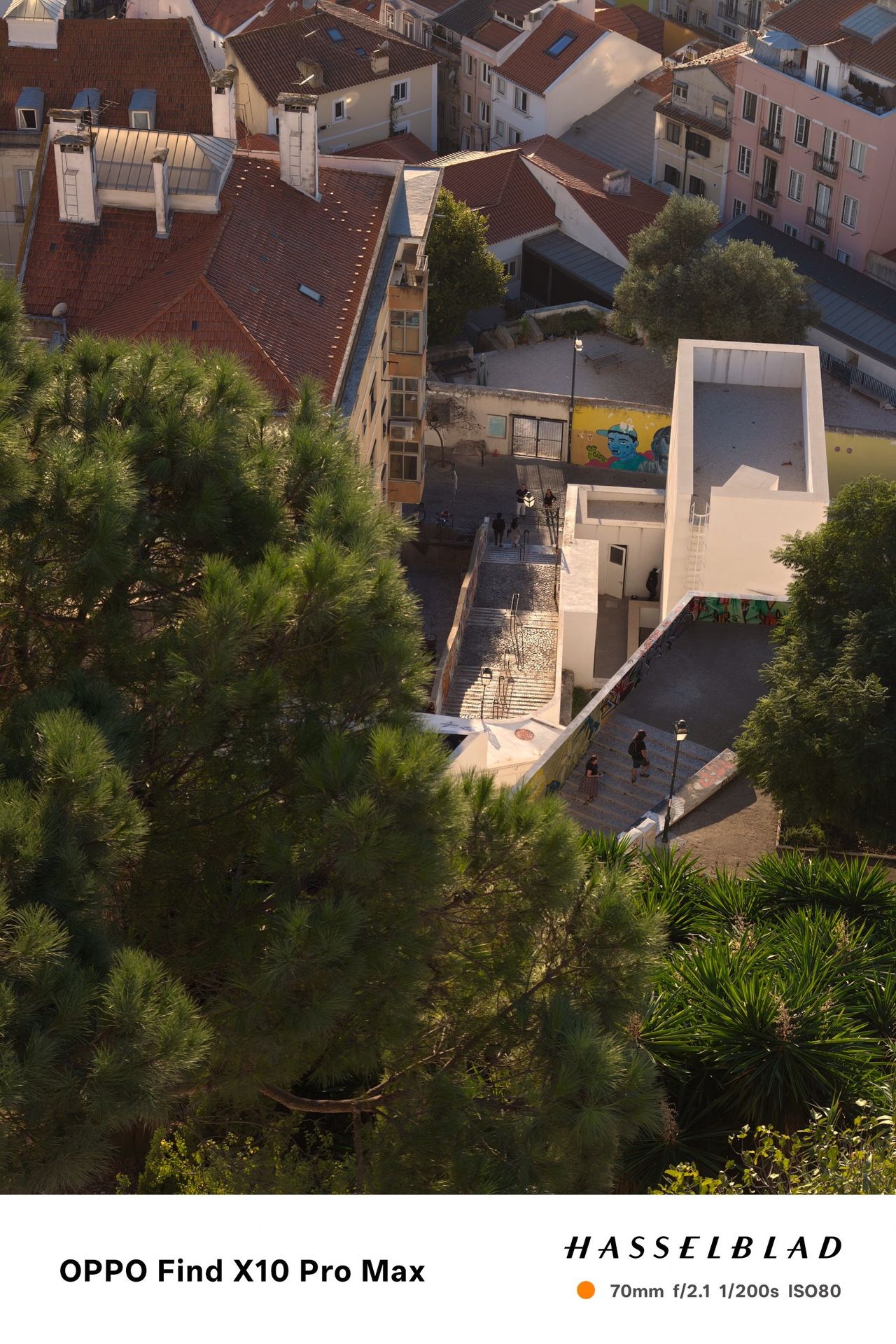 Elevated view over Lisbon rooftops, trees and a modern white building in warm evening light.