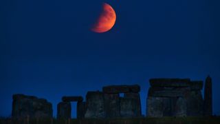 red half moon rises over stones of stonehenge that are barely visible in deep shadow. the sky is dark blue