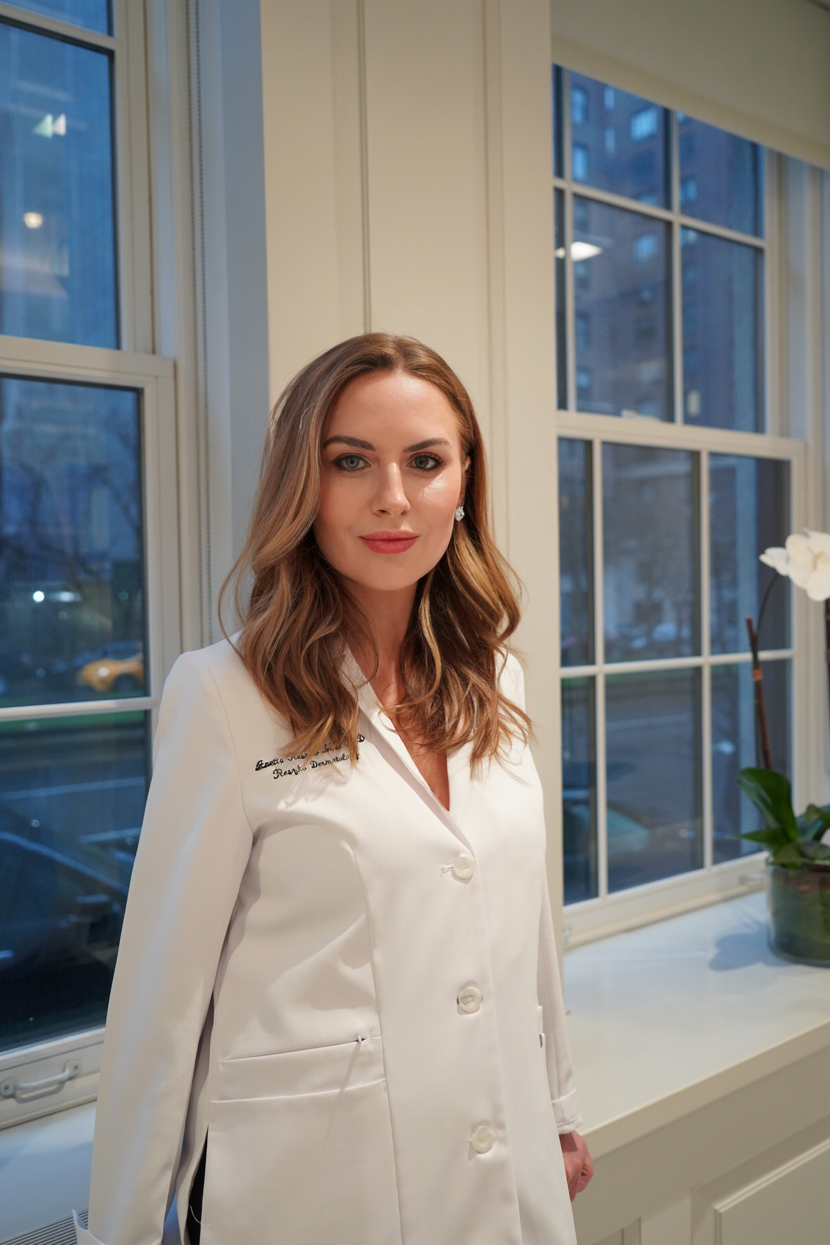 a blonde woman in a white lab coat in front of glass windows at dusk