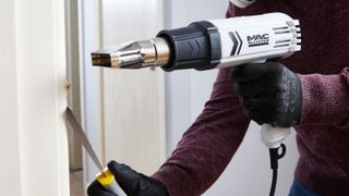 White heat gun with silver nozzle being held against painted white door frame with a paint scraper in other hand scraping paint off frame