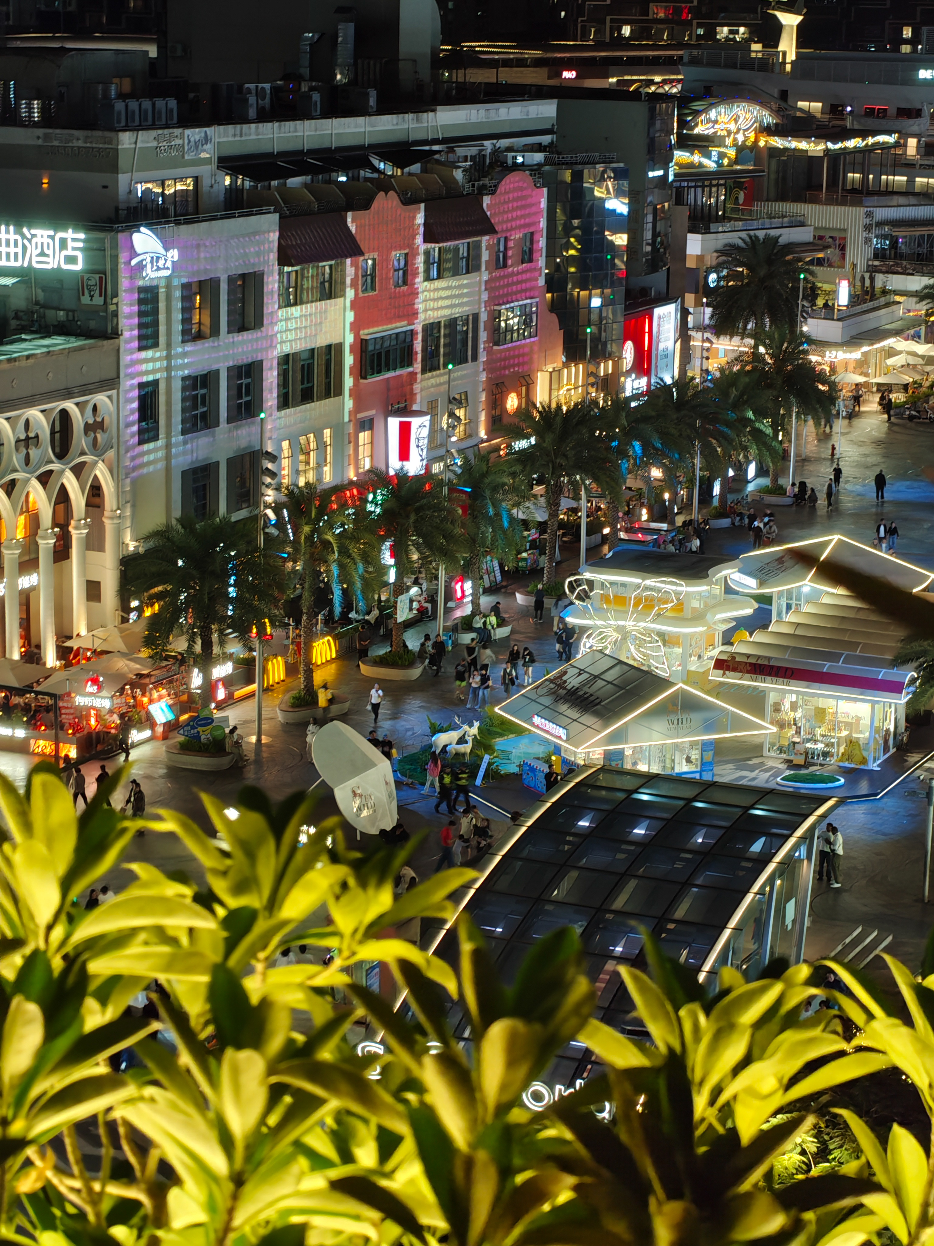 A high-angle night shot of a bustling outdoor commercial district. Buildings with illuminated storefronts and colorful facades line a pedestrian area filled with palm trees and people. Several small, brightly lit kiosks with peaked roofs are scattered throughout the plaza. Green leaves in the foreground are slightly out of focus.