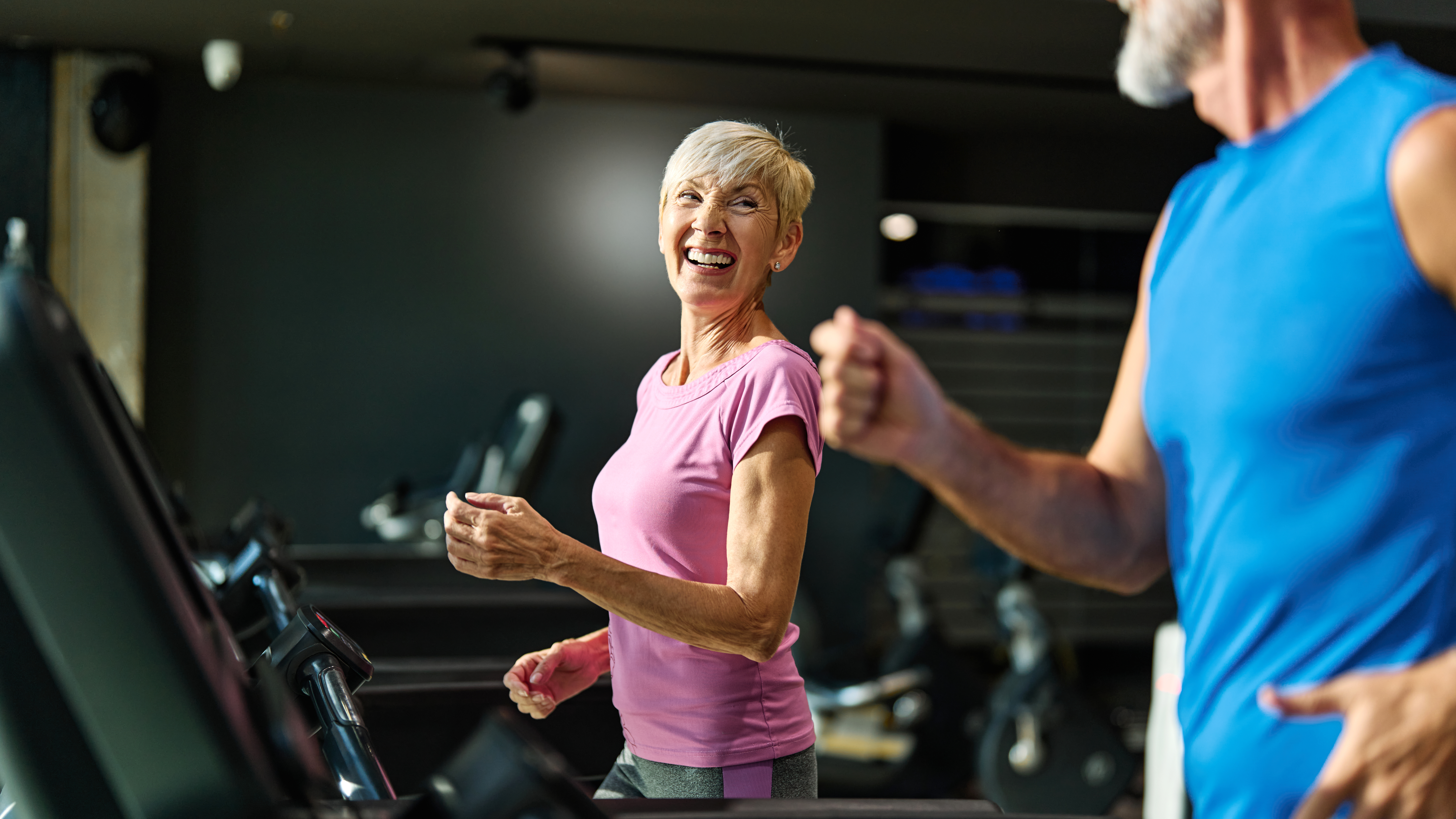 A woman walks on a treadmill and smiles at a man walking on a treadmill next to her in a gym setting.