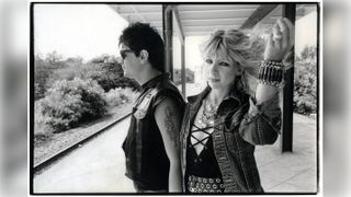 A punk couple stand on a train platform, the man in sunglasses and a leather vest, the woman in a denim jacket and bracelets, holding up her blonde hair