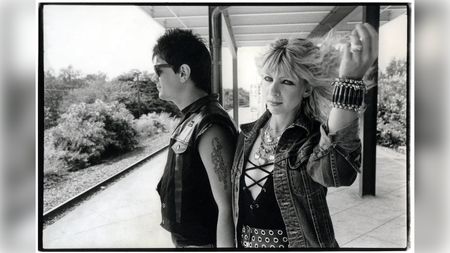 A punk couple stand on a train platform, the man in sunglasses and a leather vest, the woman in a denim jacket and bracelets, holding up her blonde hair