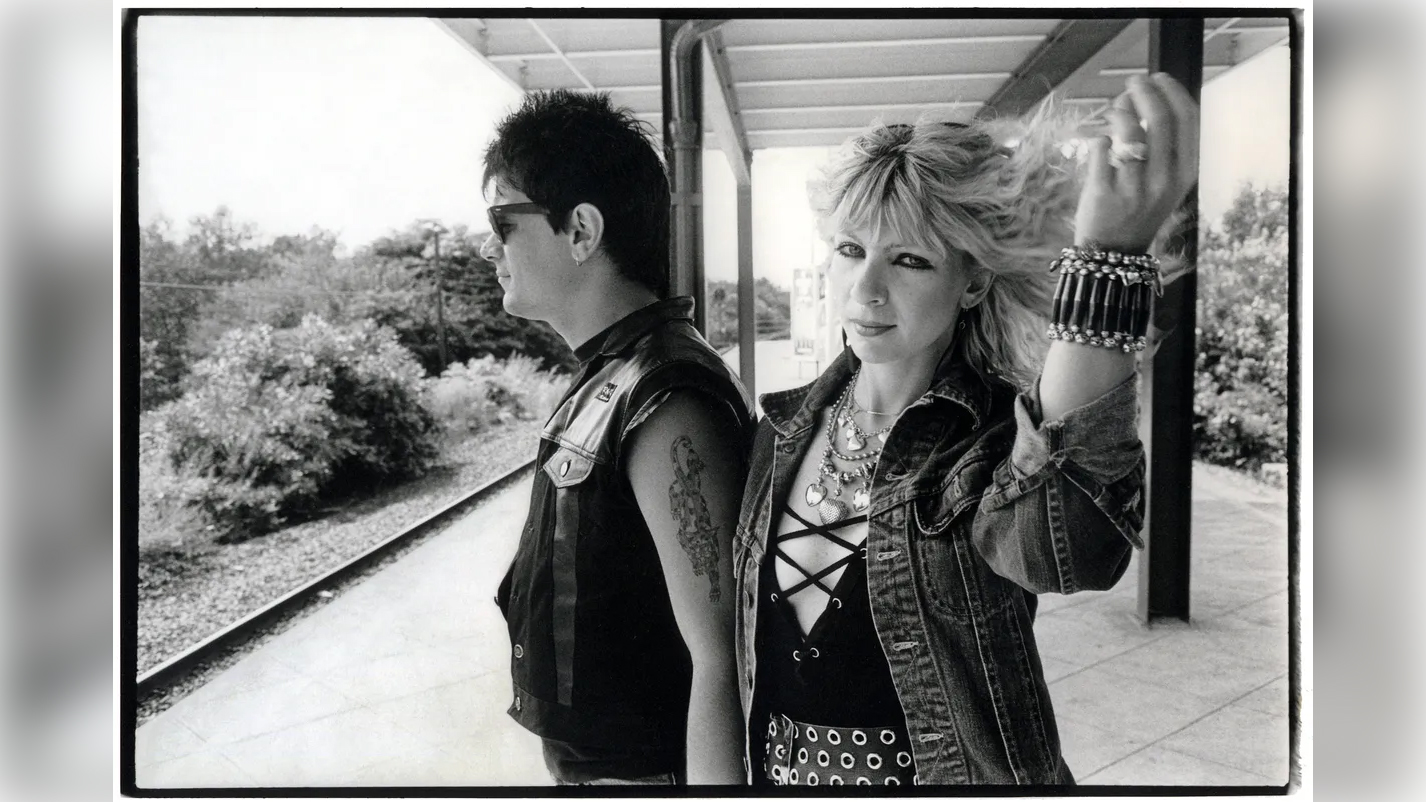 A punk couple stand on a train platform, the man in sunglasses and a leather vest, the woman in a denim jacket and bracelets, holding up her blonde hair