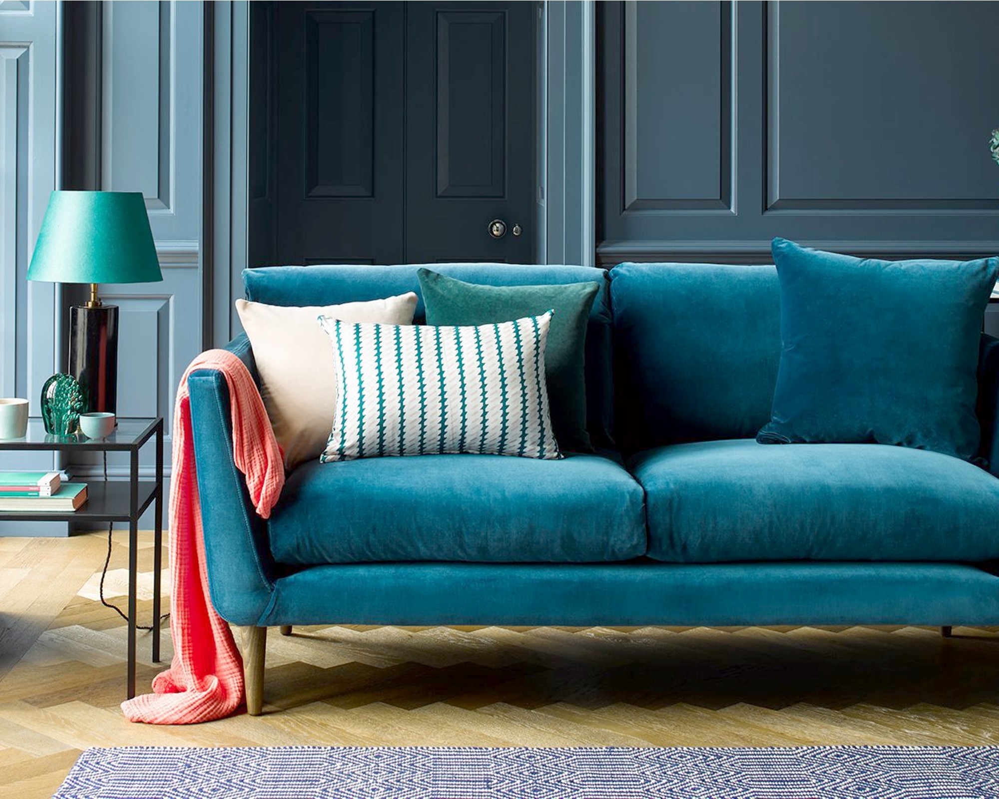 A teal blue velvet sofa with wooden legs in a living room with blue painted panelled walls
