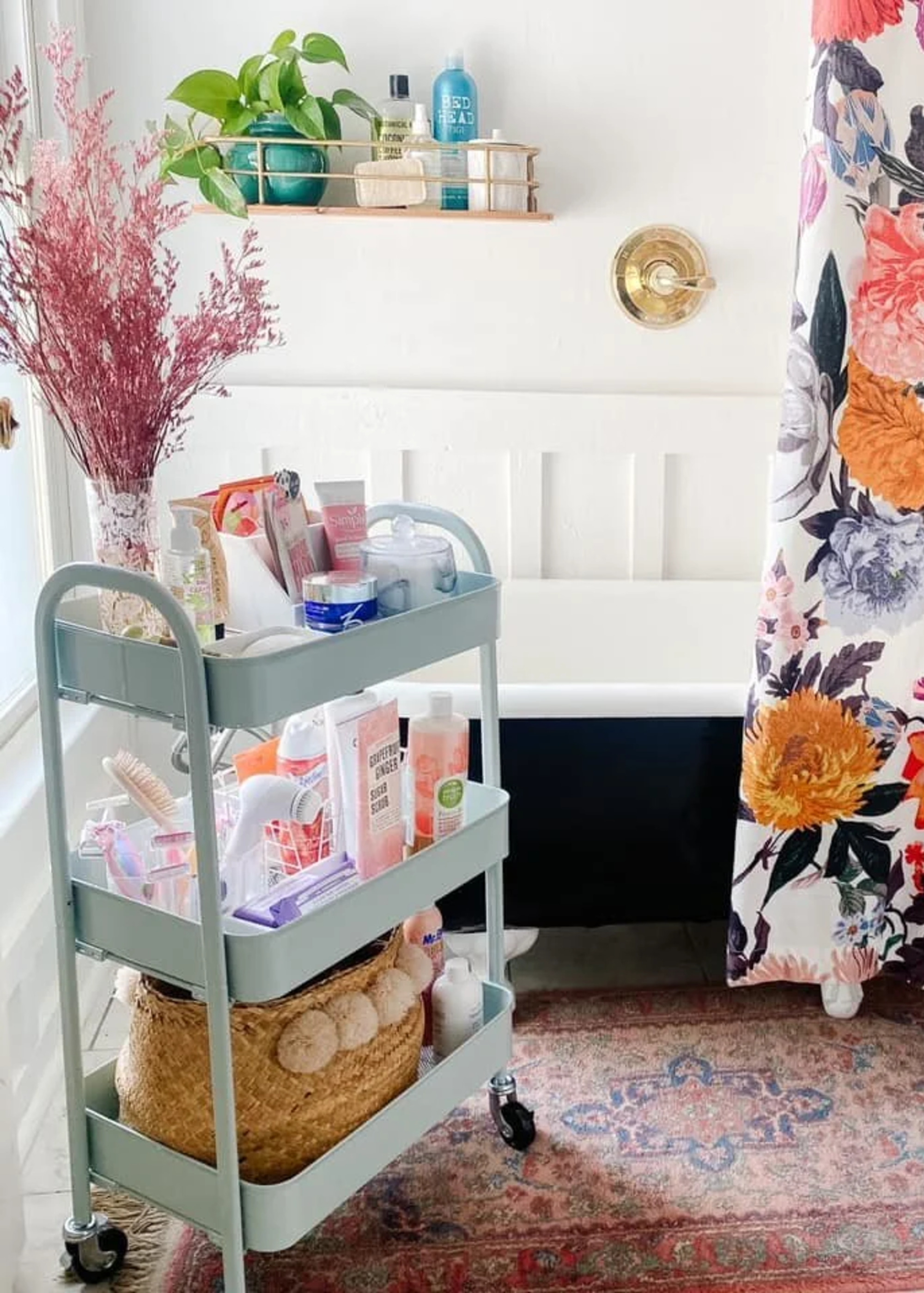 A bathroom with a black tub, a floral shower curtain, a Persian style rug and a light blue rolling cart holding toiletries