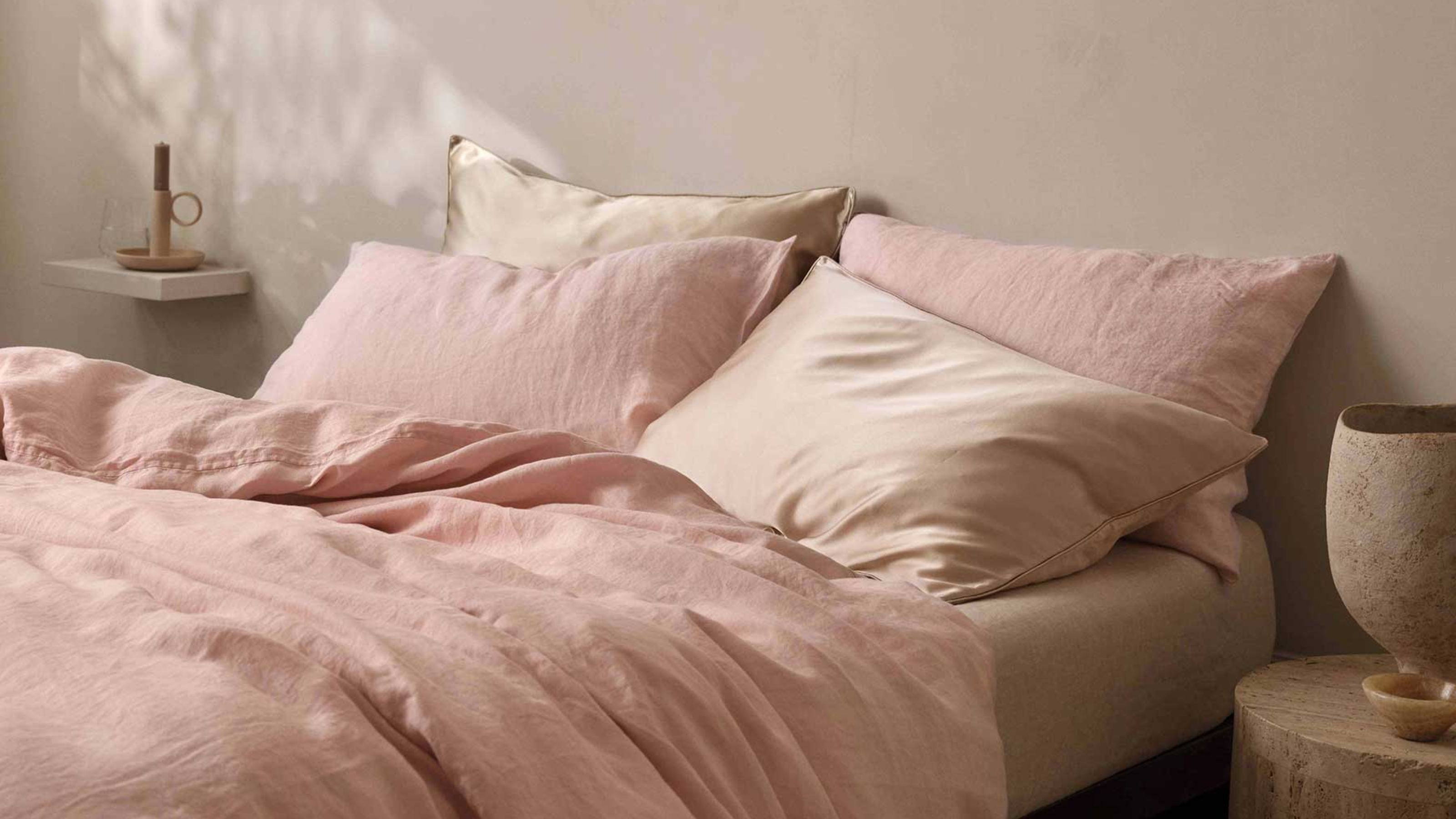 Image of a half-made bed with blush pink linen and silk bedding. The walls are beige and so are the side tables.