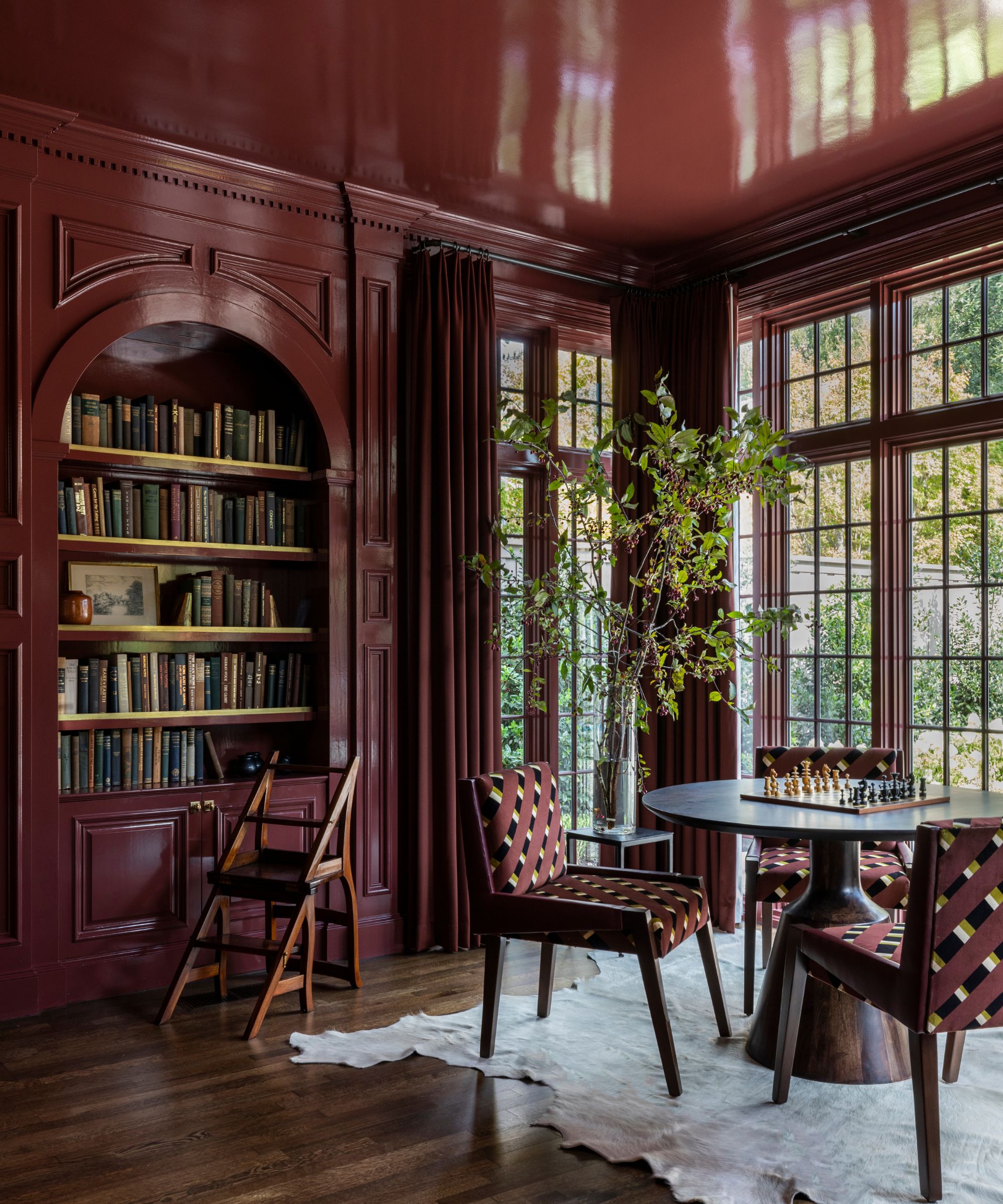 A library room with dark red color-drenched walls in a gloss finish. Floor-to-ceiling windows, built-ins, and a circular table and red chairs.