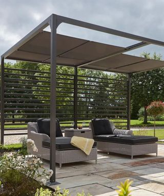 A modern pergola with retractable ceiling panels, over a patio area with plans and two brown sun loungers on it