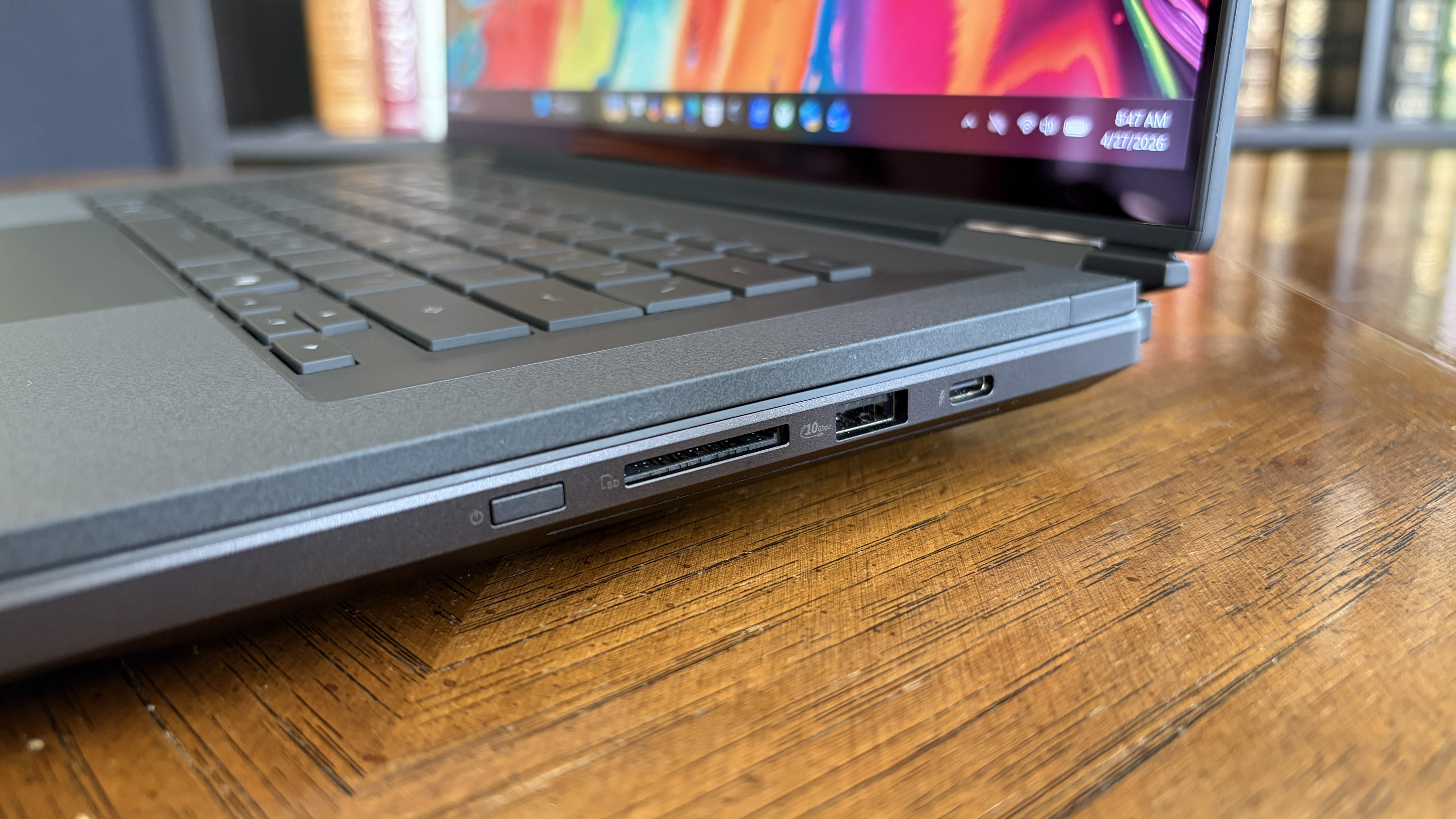 A close-up of a sleek laptop on a wooden table, showing its side ports: an SD card slot, USB port, and a USB-C port. The screen displays vibrant colors.