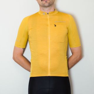 A white man in a yellow cycling jersey and black cycling shorts stands against a white wall