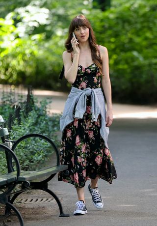 Dakota Johnson walks in a park wearing a Reformation floral print dress with Converse sneakers and a cardigan tied around her waist