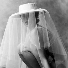 Black and white photo of woman wearing white bra, kneeling down, wearing oversize white Kyha Studios wide-brim bridal hat with veil.