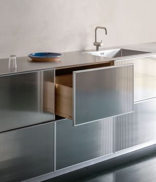 Close up of silver, semi-transparent kitchen drawers beneath a silver sink and grey worksurface with a blue painted terracotta plate and glass on top