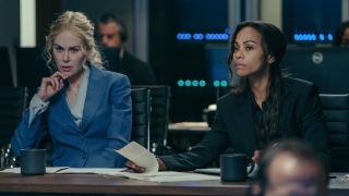 From left to right: Nicole Kidman sitting with her left elbow on her table and her hand on her chin and Zoe Saldana looking forward while lifting a paper with her left hand. 