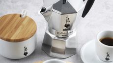 A close up of a silver Bialetti moka pot beside a lidded bowl of sugar and an espresso cup with coffee inside. 