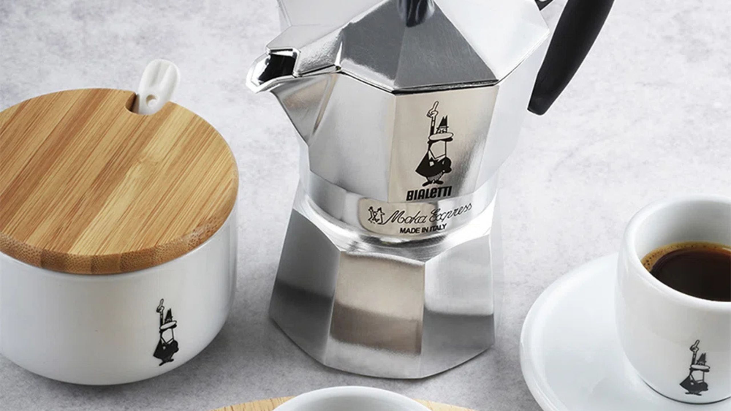 A close up of a silver Bialetti moka pot beside a lidded bowl of sugar and an espresso cup with coffee inside. 