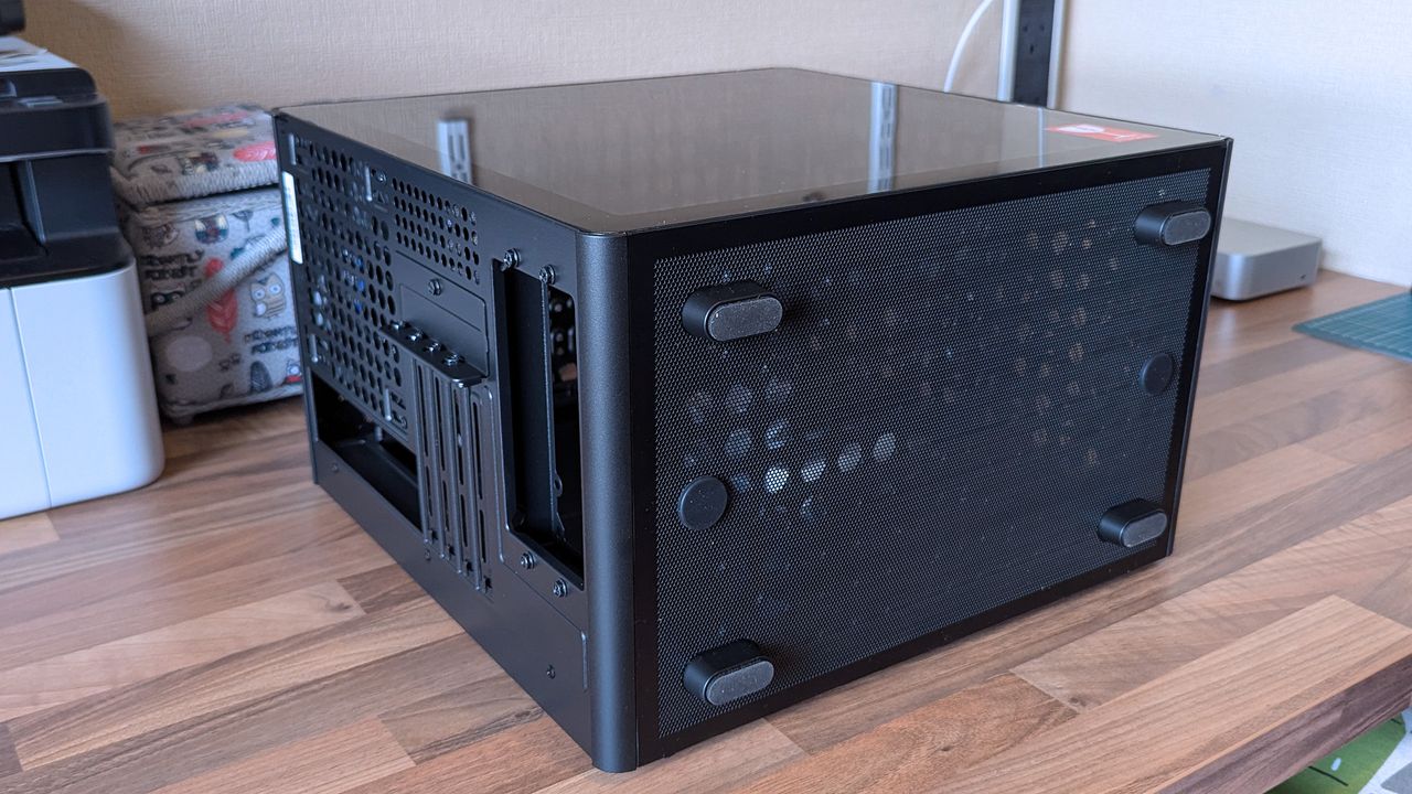 A compact Cooler Master Qube 340 case sits on a desk with a mesh bottom and tempered glass side, showing the rear expansion slots and ventilation layout.