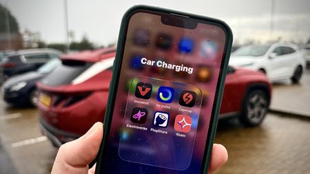 A hand holds a smartphone displaying a folder labeled "Car Charging" with six charging apps, in front of parked electric cars on a rainy day.